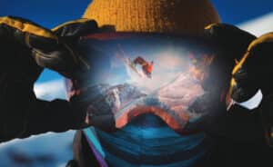 Close-up portrait of a skier in ski goggles holding on to ski goggles in the reflection of which the skier does a backflip trick. against the backdrop of snowy epic high mountains.